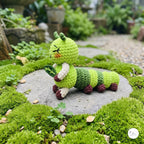 A crocheted green caterpillar toy holding a leaf, posed on a stone path surrounded by lush green moss in an outdoor garden setting.