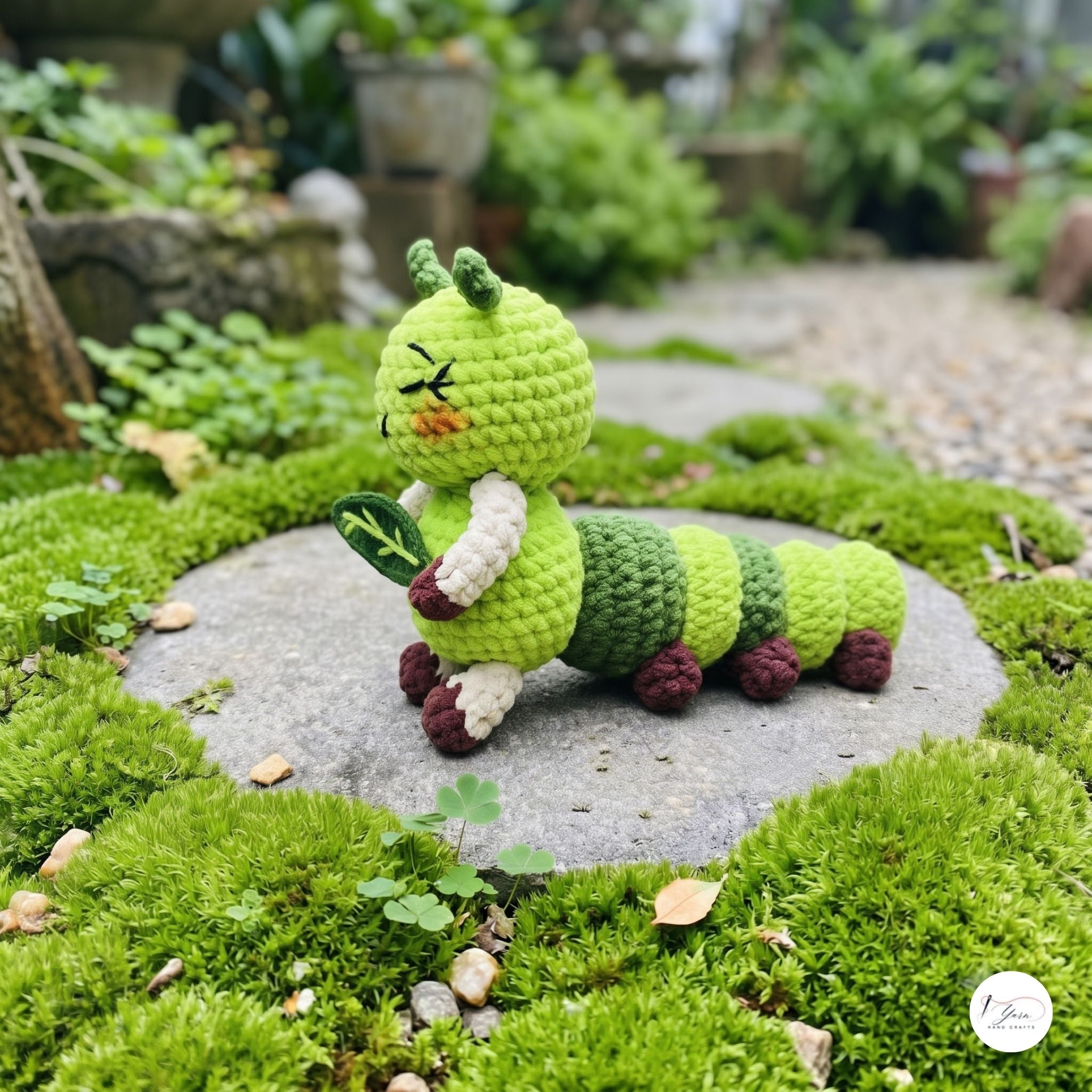 A crocheted green caterpillar toy holding a leaf, posed on a stone path surrounded by lush green moss in an outdoor garden setting.