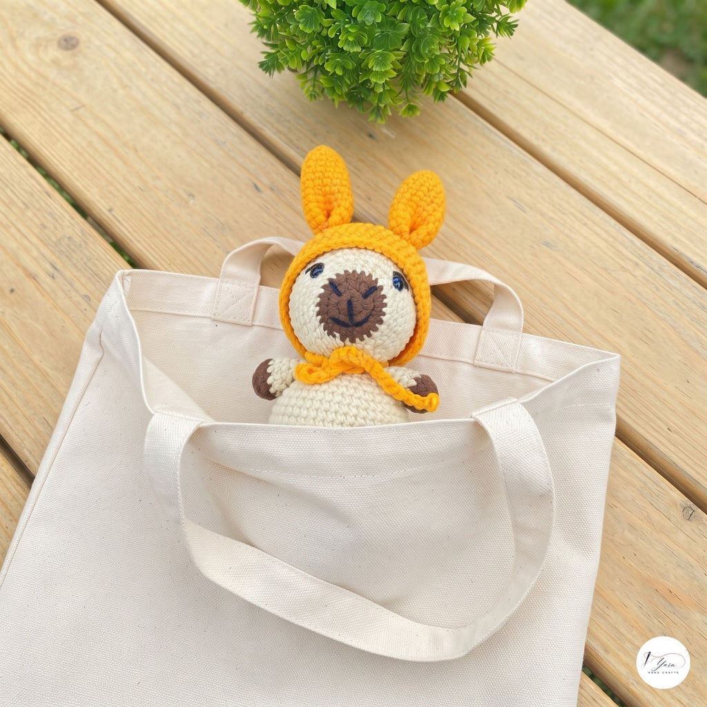 A cream canvas tote bag on a wooden table with a small crocheted llama doll peeking out from inside the bag.