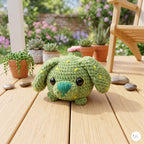 A green crocheted plush cactus dog toy with yellow spines and a pink flower on its head, sitting on a wooden deck with potted succulents in a garden background.