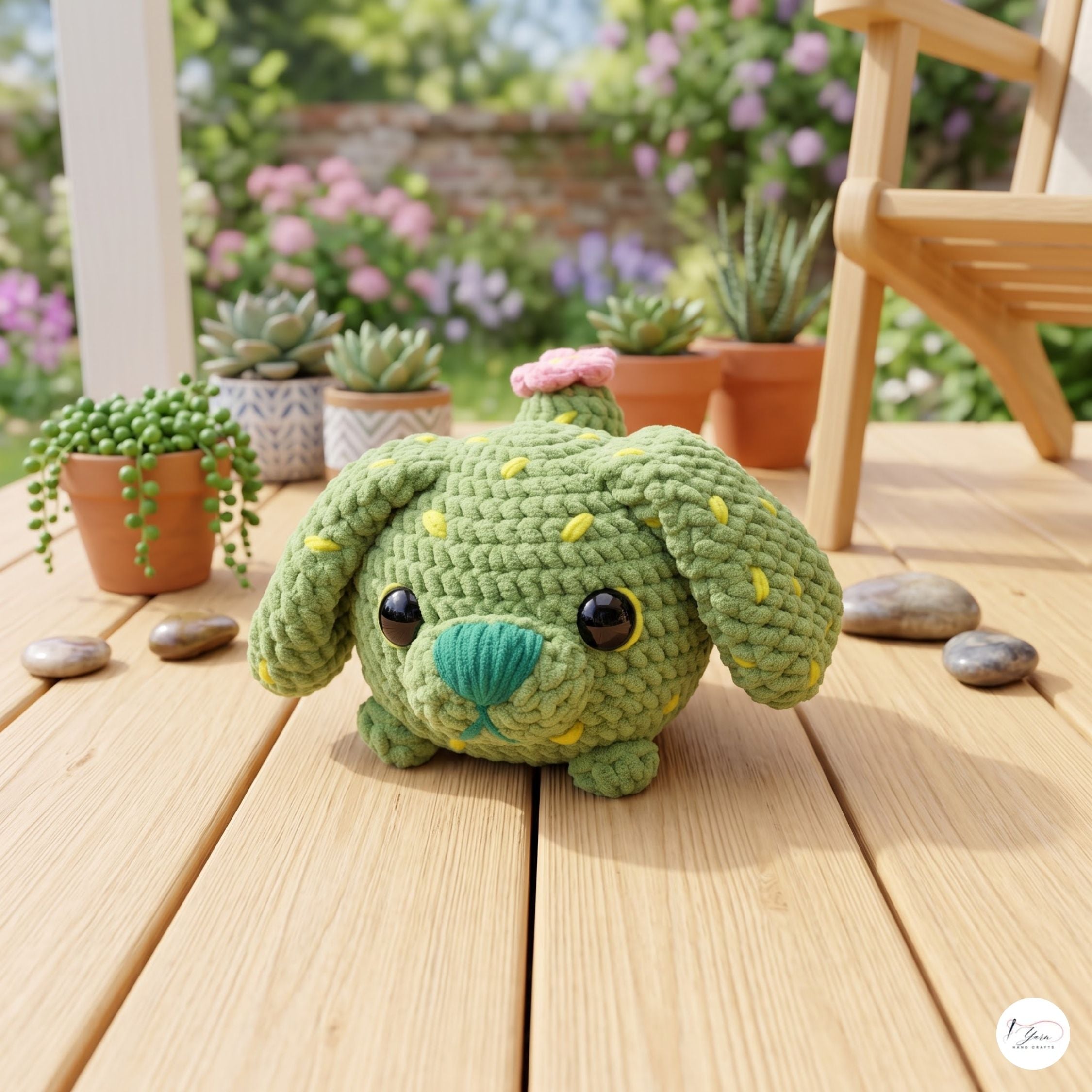 A green crocheted plush cactus dog toy with yellow spines and a pink flower on its head, sitting on a wooden deck with potted succulents in a garden background.
