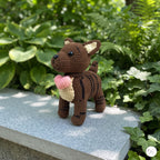 A crocheted plush dog toy with brown and black stripes, cream ears and belly patch, and a pink heart-shaped bow, standing on a gray stone ledge with green garden foliage in the background.