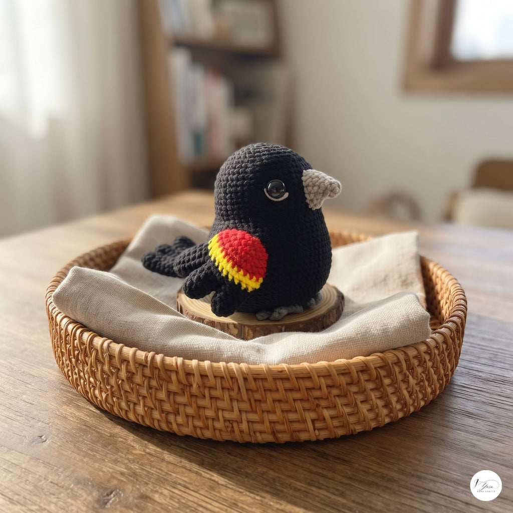 A crocheted black bird toy with a red and yellow wing and a gray beak, displayed on a small wooden slice inside a woven wicker tray lined with a beige cloth on a wooden table.