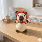 A crocheted bear plush toy wearing a red hood and scarf, sitting on a wooden table with toy blocks and yarn books in the background.