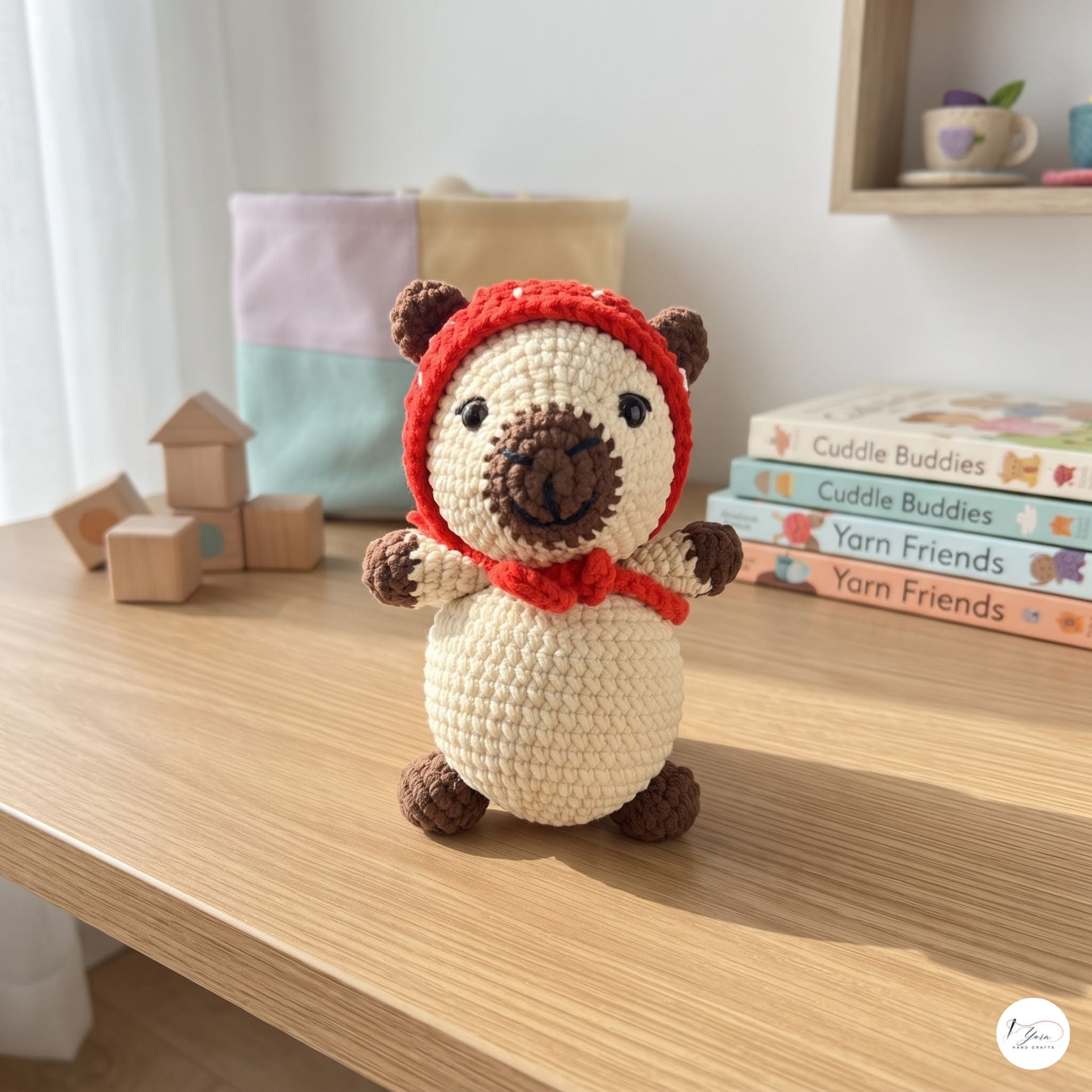 A crocheted bear plush toy wearing a red hood and scarf, sitting on a wooden table with toy blocks and yarn books in the background.