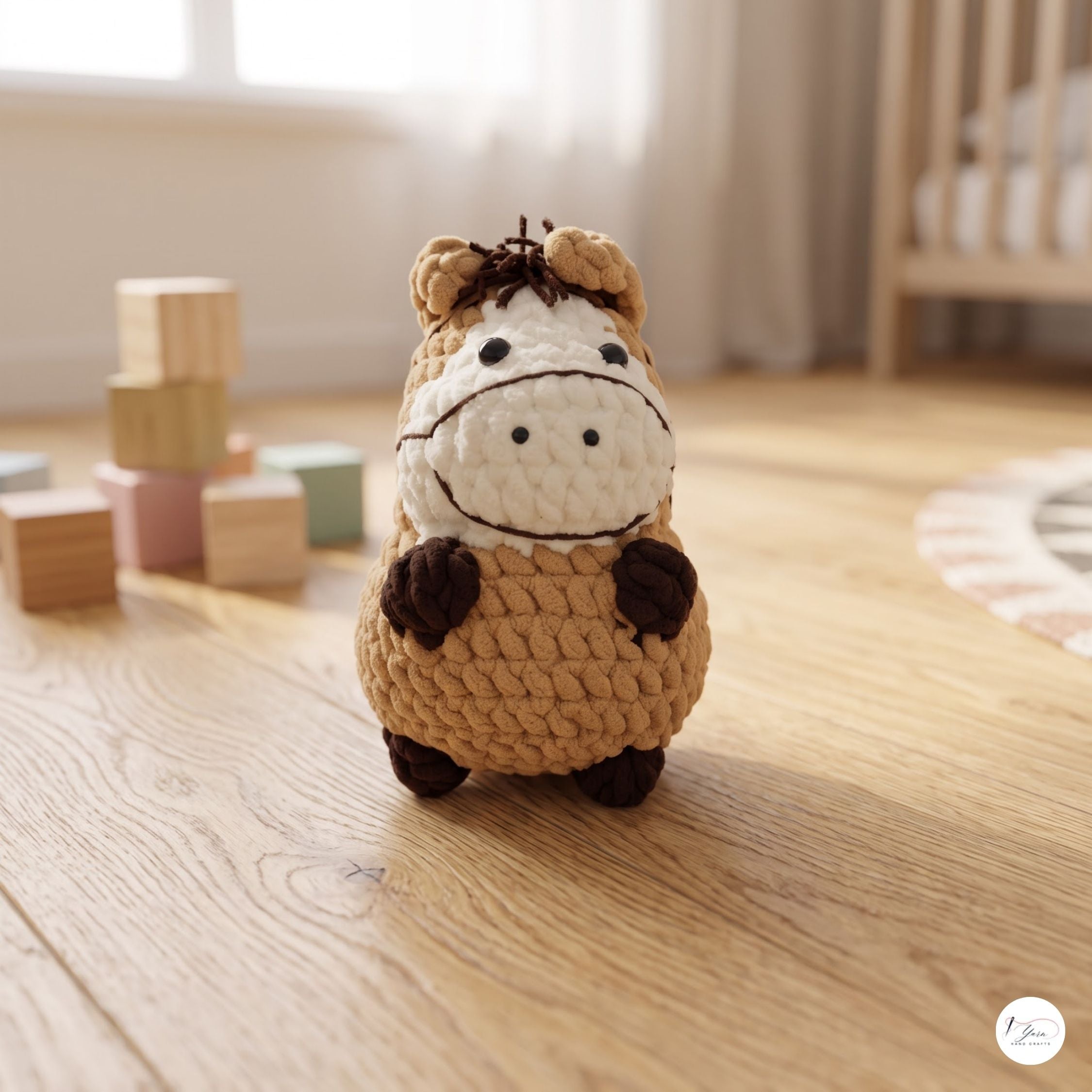 A crocheted plush toy shaped like a cow stands on a light wood floor in a softly lit nursery or playroom. The toy has a tan body, a cream face, and dark brown accents on the hooves, ears, and muzzle.