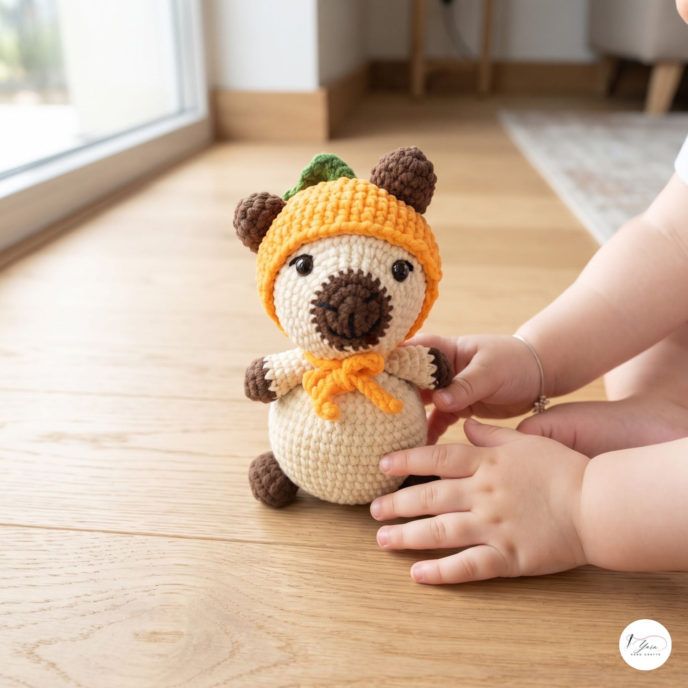 A handmade crocheted plush bear toy wearing a yellow-orange hat and bow tie, sitting on a light wooden floor with a child's hands gently touching it. A small circular 'The Amigurumi Shop' logo is visible in the bottom right corner.