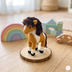 Crocheted amigurumi horse toy standing on a round wood slice, photographed on a wooden floor with soft-focus toys and a rainbow in the background.