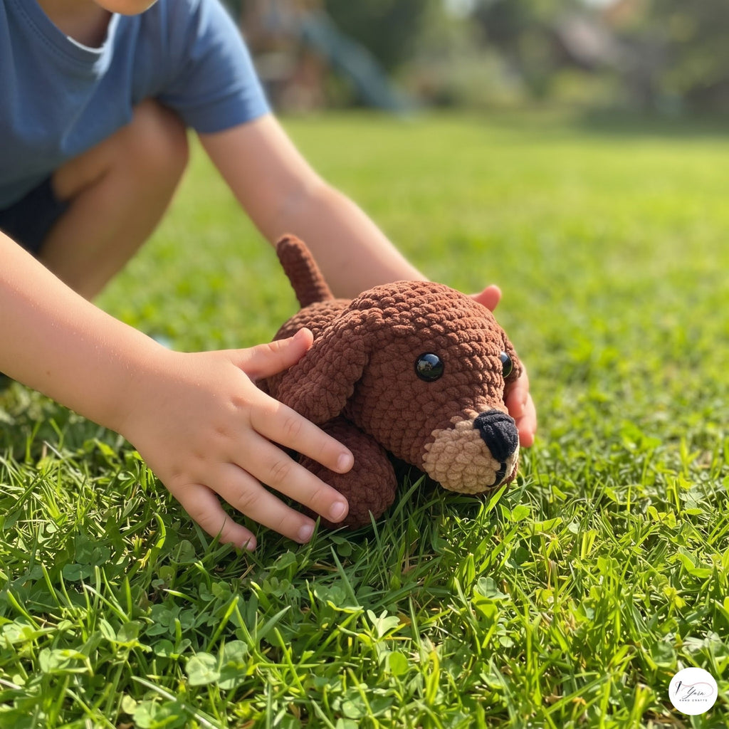 A child's hands gently holding a small brown crocheted dog toy on a green grass lawn.