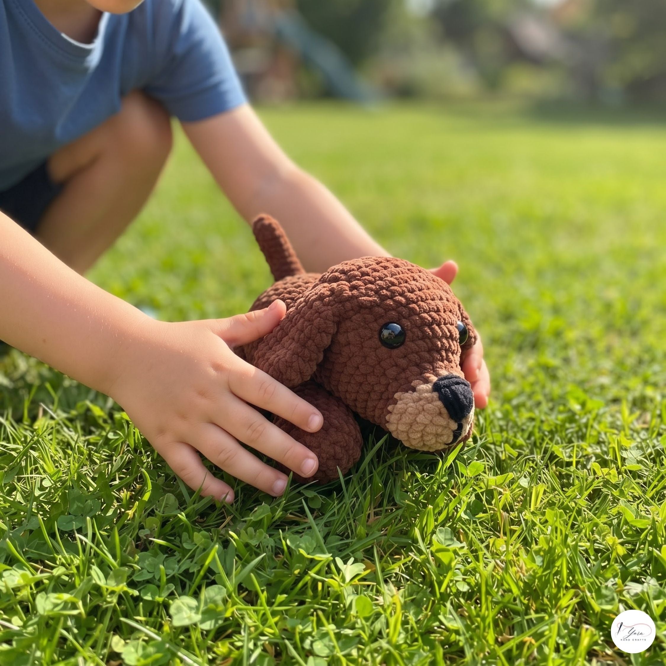 A child's hands gently holding a small brown crocheted dog toy on a green grass lawn.