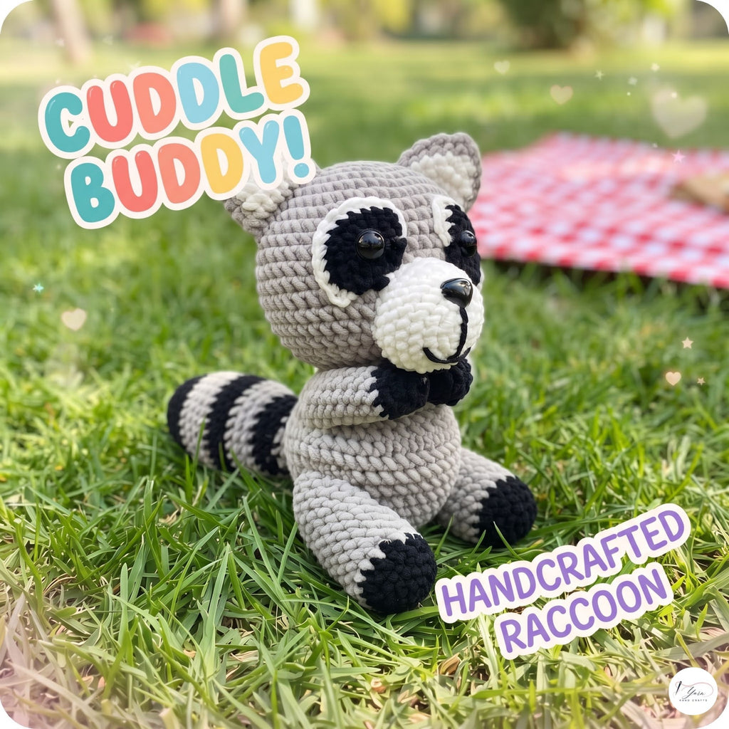 A crocheted raccoon toy sitting on green grass outdoors, with colorful text overlays reading 'CUDDLE BUDDY!' and 'HANDCRAFTED RACCOON'. A blurred red-and-white picnic blanket is visible in the background.