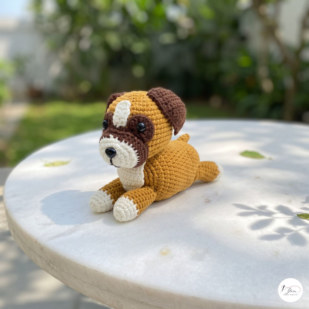 A crocheted dog toy sits on a round white table outdoors, with a blurred garden background. The toy is made of brown, cream, and white yarn, with dark brown ears, muzzle, and paws, and a cream-colored chest, paws, and snout.