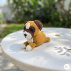 A crocheted dog toy sits on a round white table outdoors, with a blurred garden background. The toy is made of brown, cream, and white yarn, with dark brown ears, muzzle, and paws, and a cream-colored chest, paws, and snout.