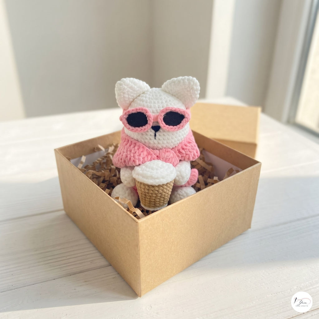 A small crocheted plush toy with a white body, pink scarf, and pink sunglasses sits inside a brown kraft gift box filled with crinkle paper. The plush holds a waffle cone.