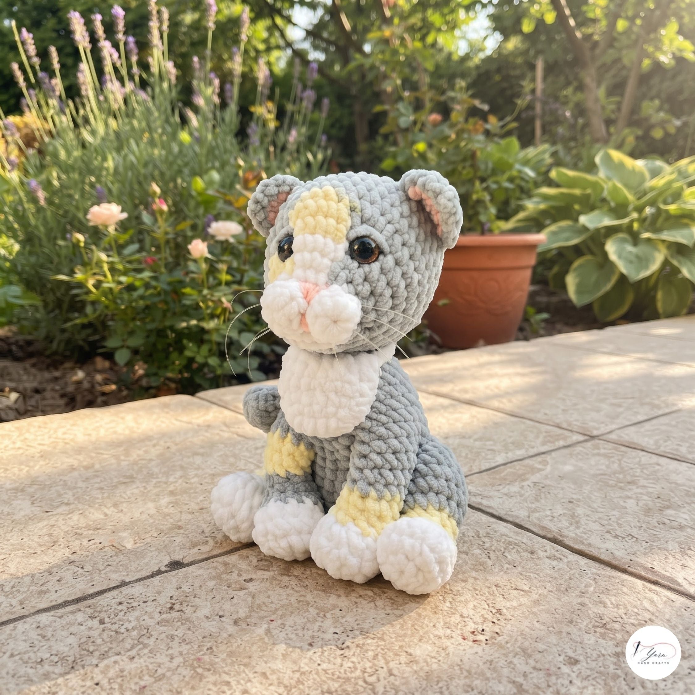 A crocheted cat toy sits on a stone patio in a garden setting. The toy is gray with white and pale yellow accents, featuring black bead eyes and a pink stitched nose.