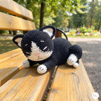 A crocheted black and white cat toy lying on a wooden park bench with a blurred outdoor garden background.