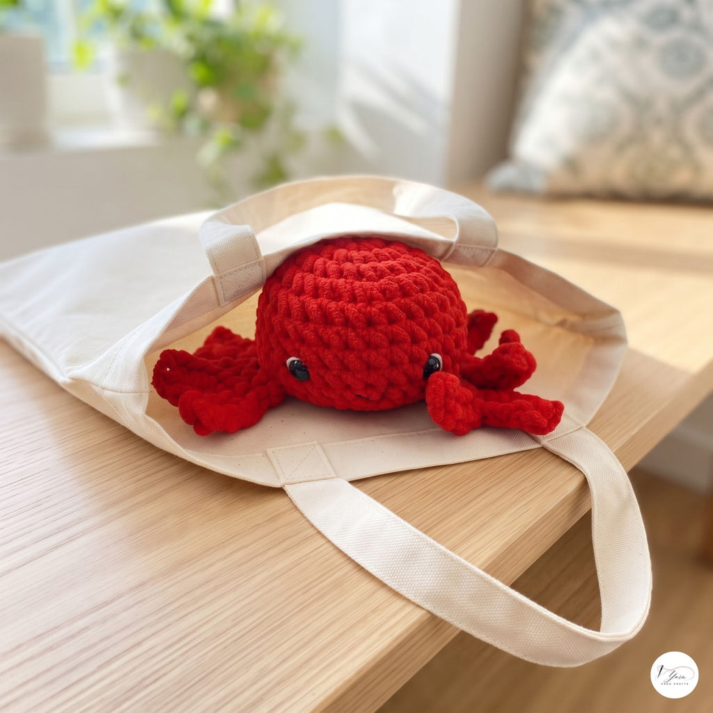 A red crocheted octopus toy peeking out from a cream canvas tote bag, placed on a light wood table in a softly lit indoor setting.