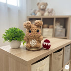 A crocheted plush bunny toy in tan and cream sits on a light wood shelf beside a small potted green plant and a red-and-white knitted egg, with a softly blurred nursery room background.