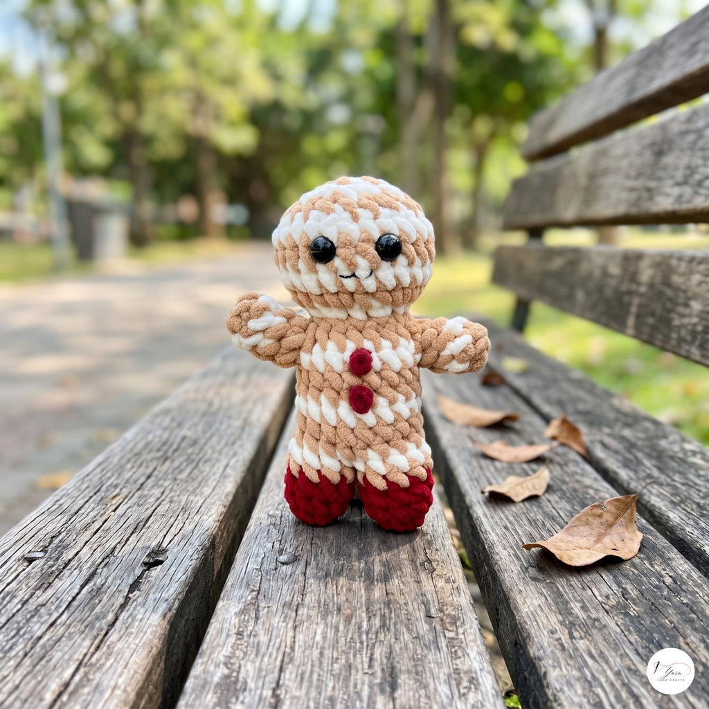 A small crocheted gingerbread person toy stands on a weathered wooden park bench with blurred trees in the background.