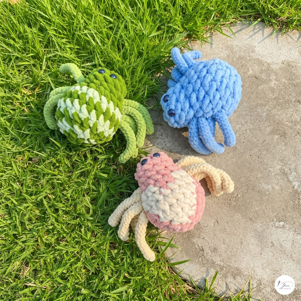 Three crocheted crab toys in green, blue, and pink-and-cream are placed on grass and concrete outdoors.
