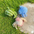 Three crocheted crab toys in green, blue, and pink-and-cream are placed on grass and concrete outdoors.