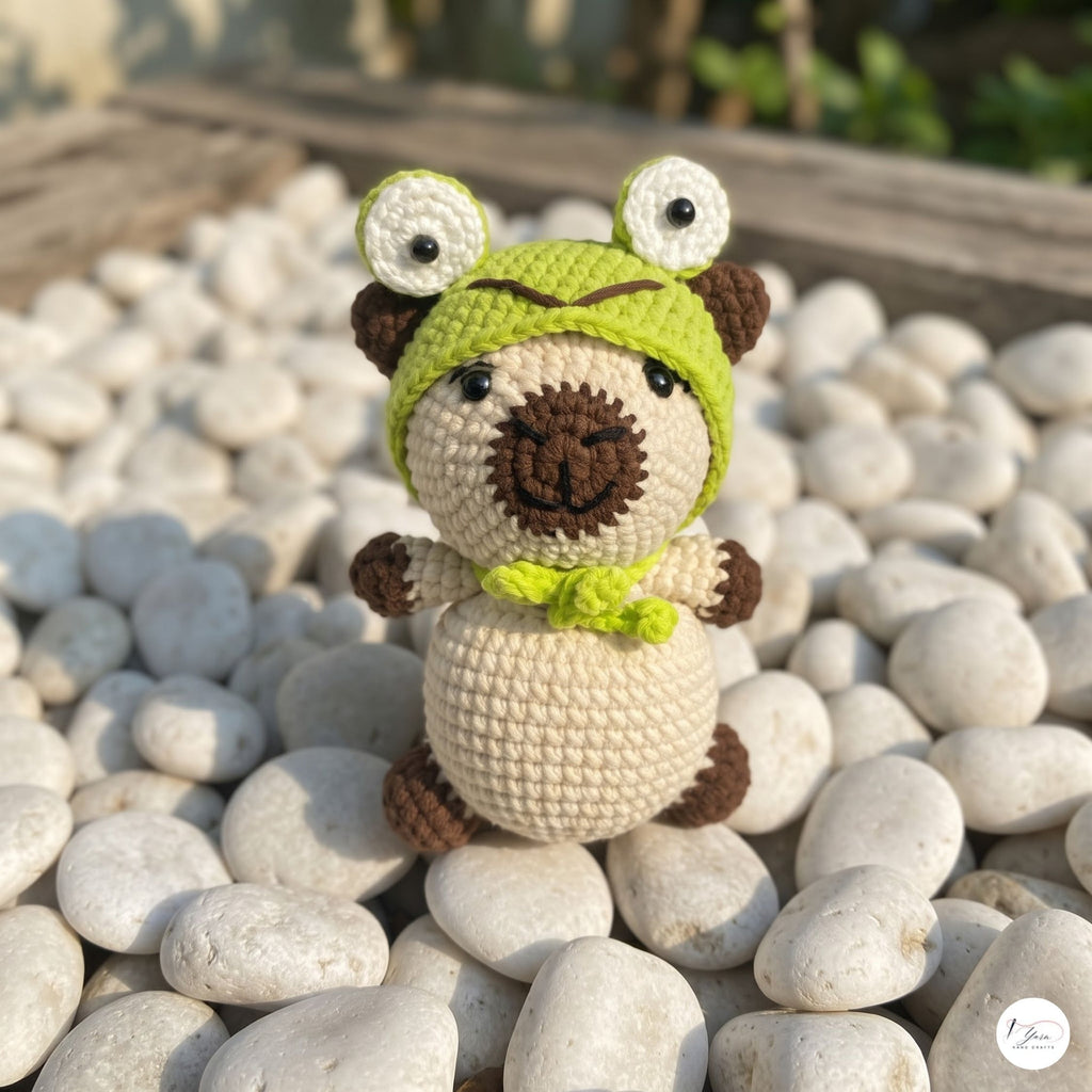 A crocheted plush toy resembling a frog or animal character, standing on white pebbles outdoors. The toy has a cream body, brown limbs and snout, and a bright green hat with large white eyes.
