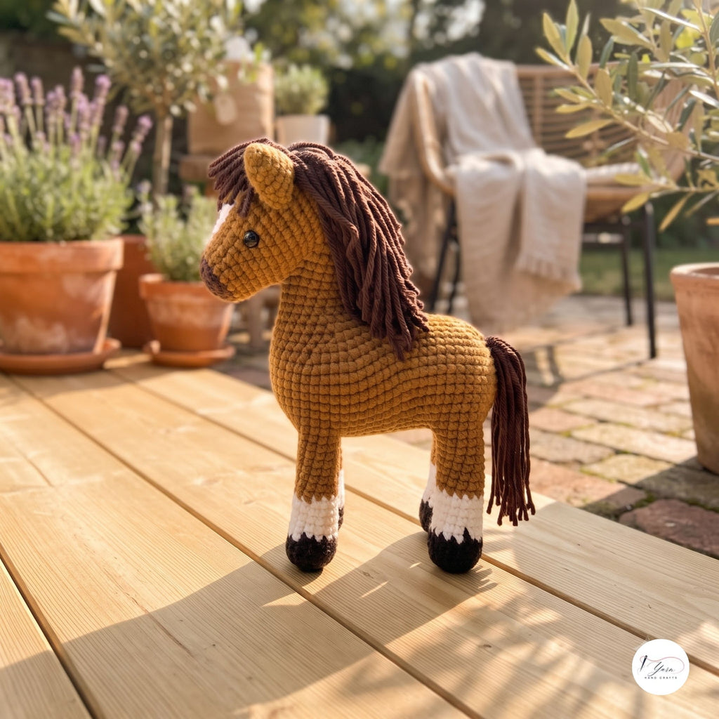 A crocheted toy horse standing on a wooden deck in a sunny outdoor garden setting with potted plants and a chair in the background.