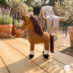 A crocheted toy horse standing on a wooden deck in a sunny outdoor garden setting with potted plants and a chair in the background.