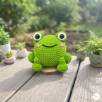 A green crocheted frog toy with large white eyes, pink cheeks, and a black stitched smile, sitting on a wooden table outdoors with blurred potted plants in the background.