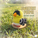 A crocheted goldfinch toy labeled 'SWEETEST PEEPS! CROCHET GOLDIE' sits on a small wooden slice in a grassy outdoor setting with warm sunlight and bokeh.