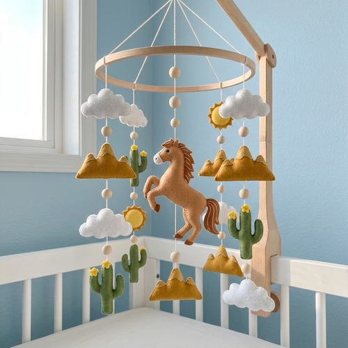 A wooden baby crib mobile hanging in a nursery, featuring a tan horse, white clouds, green cacti, and mustard-yellow mountains with small yellow flowers. The mobile is mounted on a light wood crib rail against a pale blue wall.