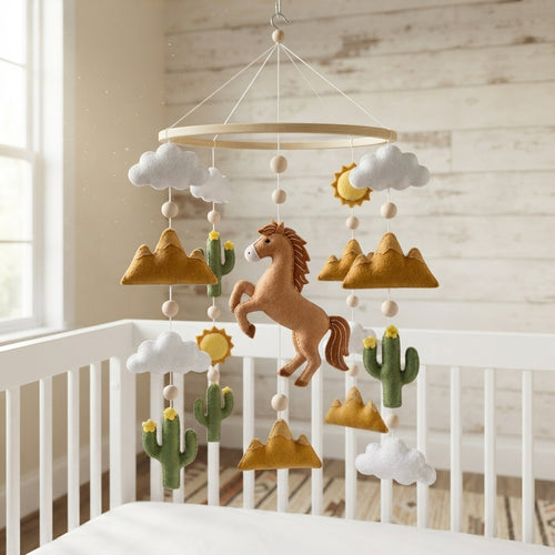 A felt baby mobile with a brown horse, cacti, clouds, and suns hanging from a wooden ring above a white crib in a softly lit nursery.