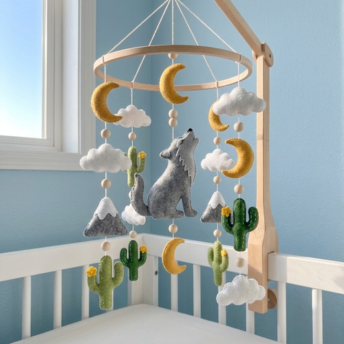 A wooden baby crib mobile hanging in a nursery, featuring a gray felt wolf, yellow crescent moons, green cacti, white clouds, and gray snowy mountains.