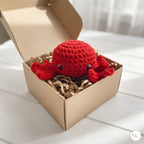 A small crocheted red octopus toy sits inside an open kraft paper box filled with brown paper shred, placed on a light wood surface.