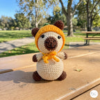 A small crocheted plush bear toy sits on a wooden picnic table outdoors in a sunny park. The toy has a cream body, dark brown ears, nose, and feet, and wears a bright orange-yellow bonnet with a small green accent and matching orange-yellow scarf.