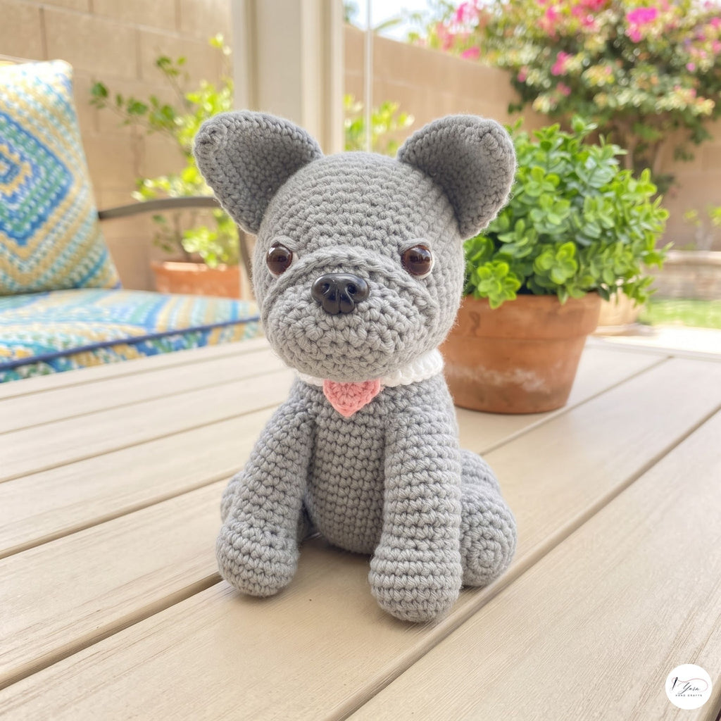 A gray crocheted dog toy with a white collar and pink heart-shaped tag, sitting on a wooden deck table with potted plants and patio furniture in the background.