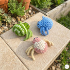 Three crocheted crab toys in green-and-white, blue, and pink-and-cream are arranged on a light stone patio outdoors with plants and gravel visible in the background.