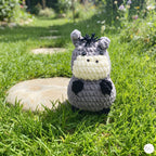 A small crocheted plush toy shaped like a cartoon donkey or hippo sits on green grass outdoors, with a stone path and blurred garden background.