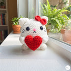A white crocheted cat plush toy holding a red heart, sitting on a white windowsill with green potted plants in the background.