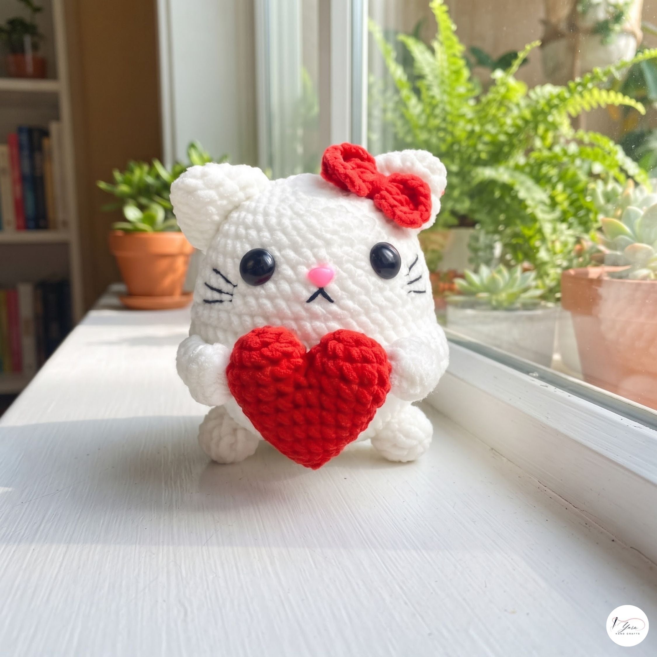 A white crocheted cat plush toy holding a red heart, sitting on a white windowsill with green potted plants in the background.