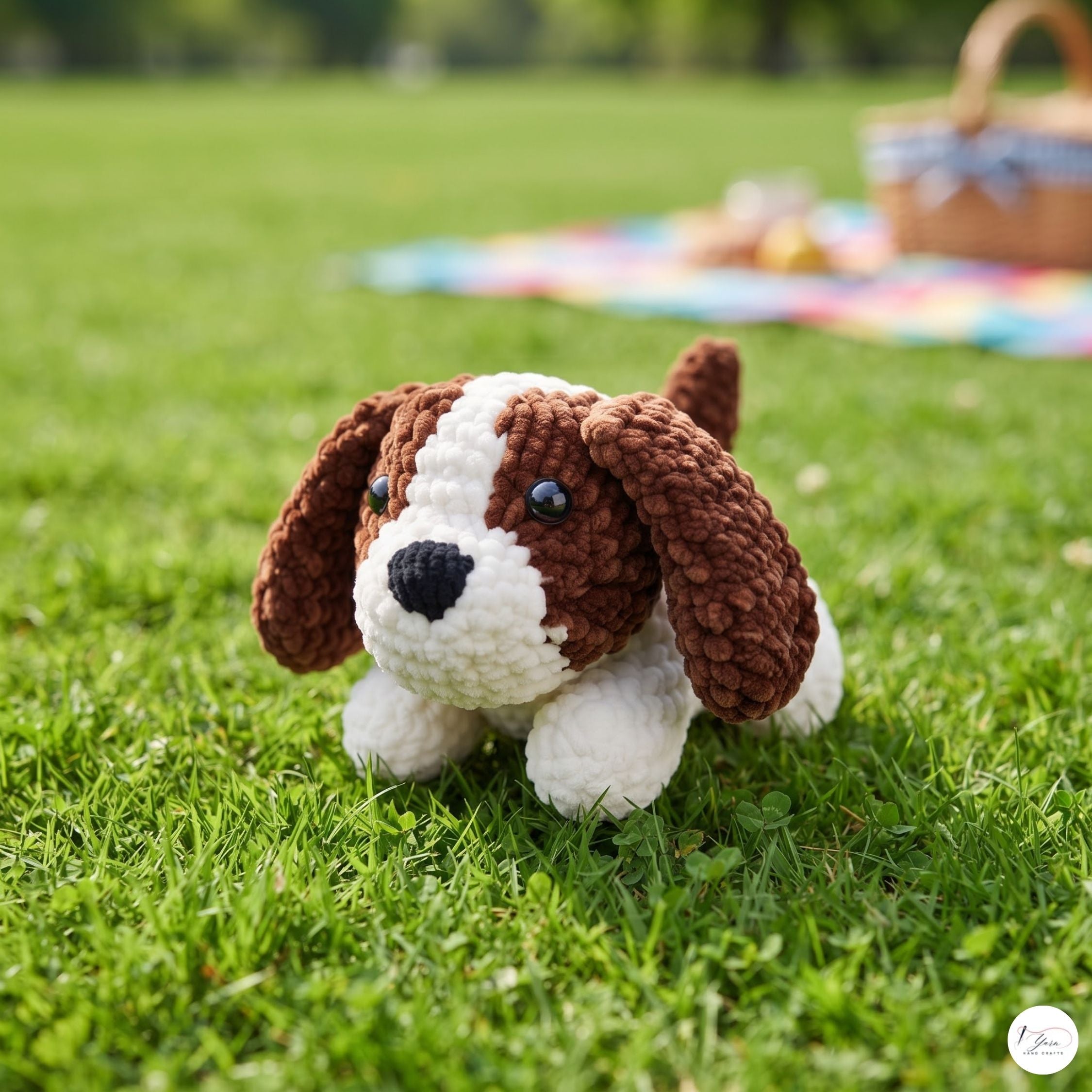 A crocheted dog toy with brown and white yarn sits on a green grass lawn, with a blurred picnic blanket and basket in the background.