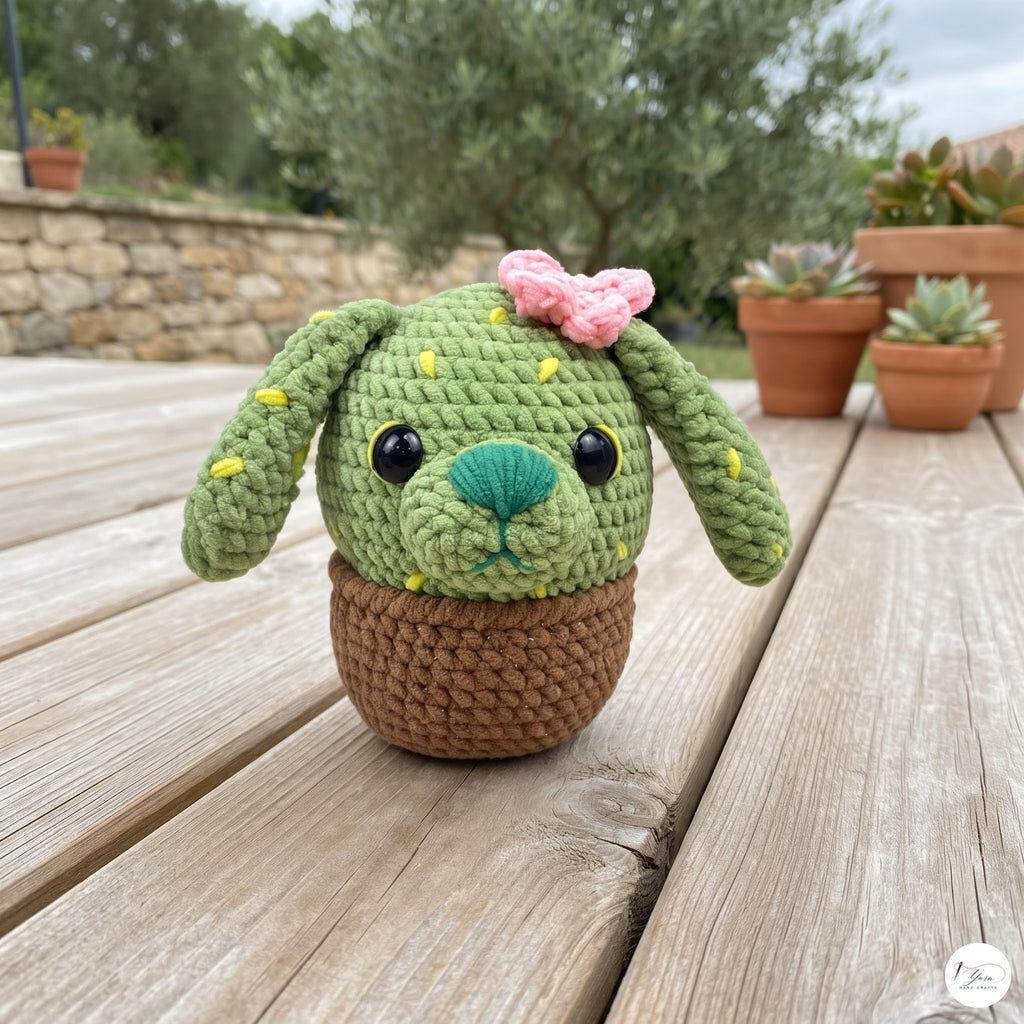 A handmade crocheted cactus dog toy sits on a wooden deck with potted succulents in the background. The toy features a green cactus head with floppy ears, yellow spines, a pink bow, a teal nose, and a brown pot base.