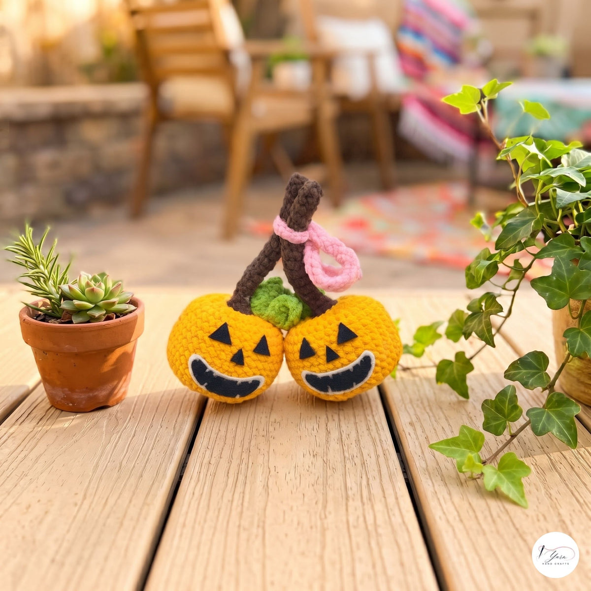 Two small yellow crocheted jack-o'-lantern toys with black embroidered faces and brown stems, tied together with a pink loop, displayed on a wooden table in an outdoor patio setting with potted plants.