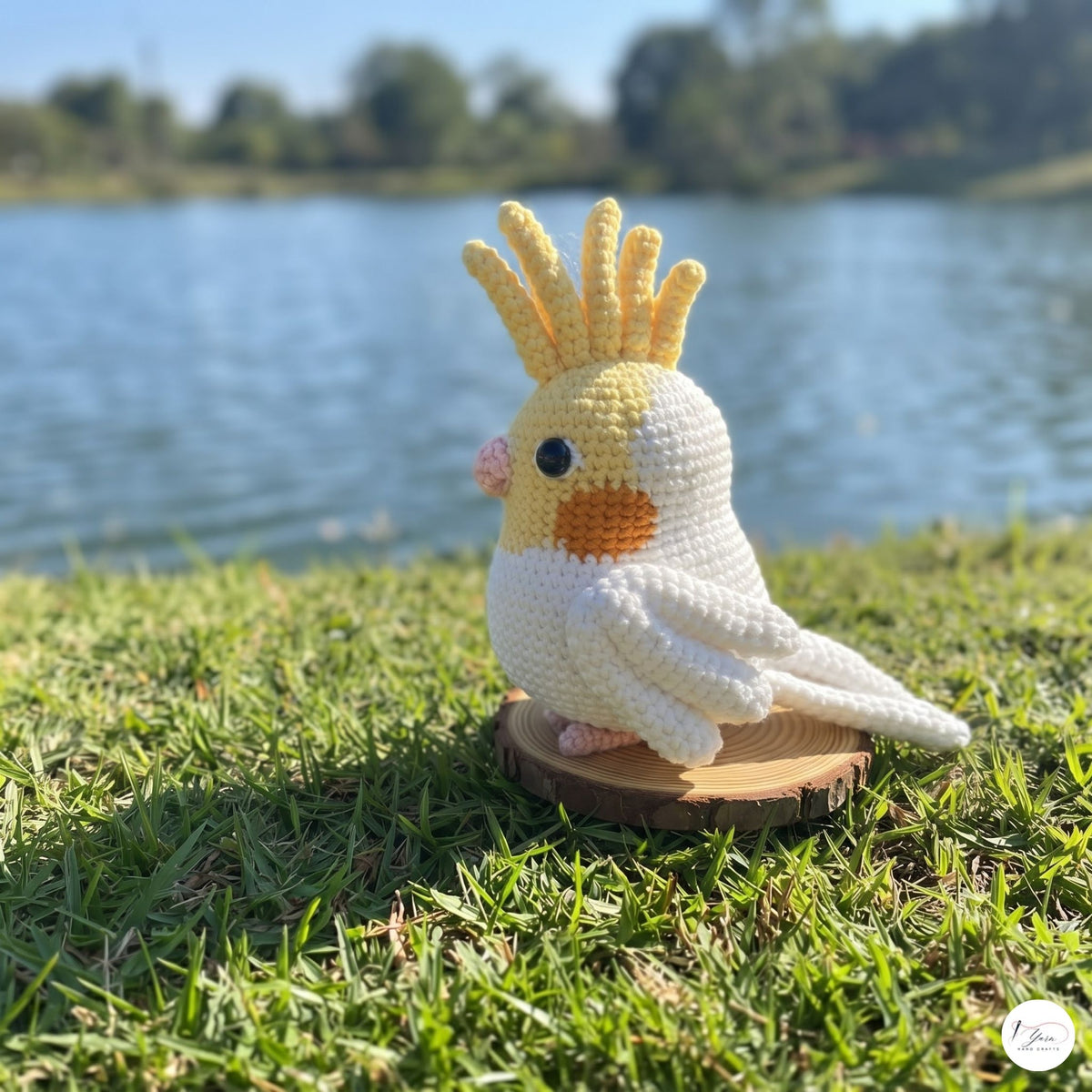 A crocheted cockatiel bird figurine in white and yellow, standing on a small wooden slice outdoors on green grass with a lake and trees in the background.