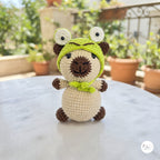 A crocheted plush toy resembling a pug wearing a green frog hood, sitting on a light gray marble table with a blurred outdoor garden background.