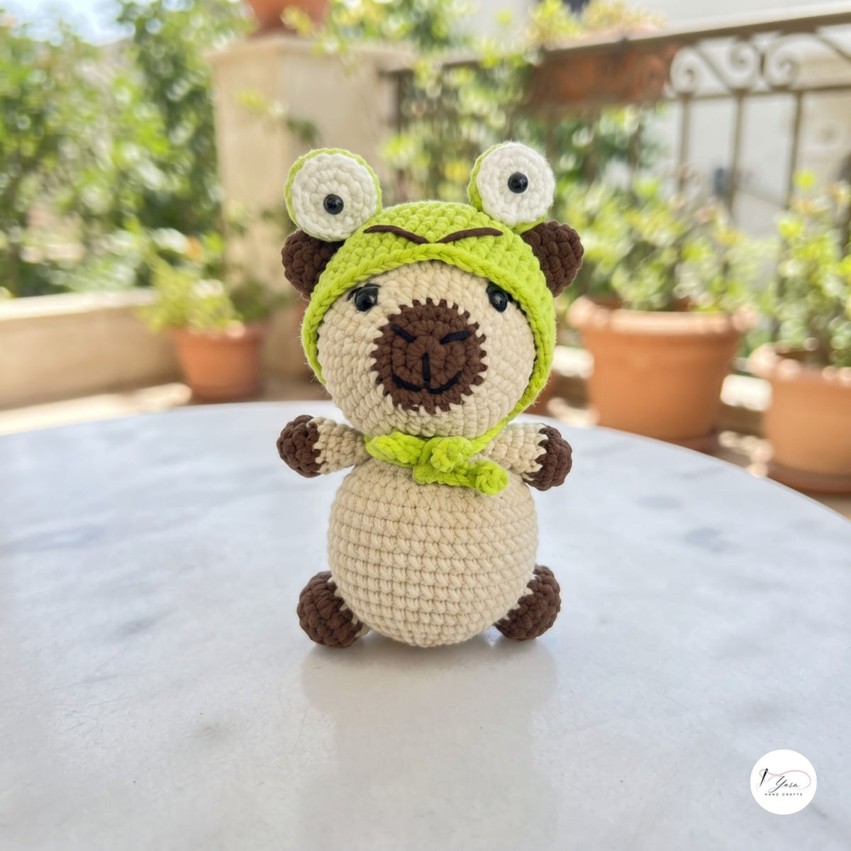 A crocheted plush toy resembling a pug wearing a green frog hood, sitting on a light gray marble table with a blurred outdoor garden background.