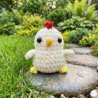 A small crocheted chick toy sits on a stone in a garden lawn. The chick is off-white with yellow feet and beak, black bead eyes, and a red heart-shaped comb.