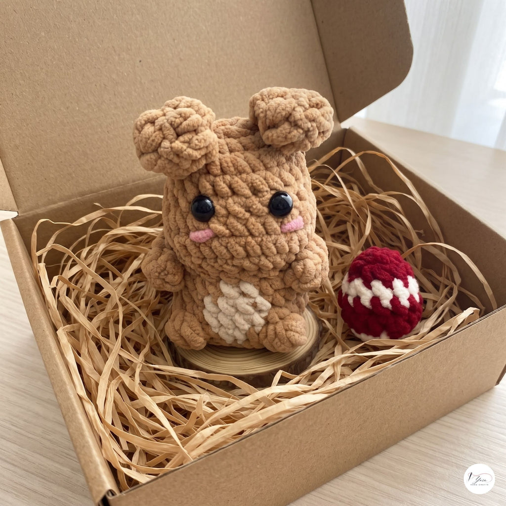 A crocheted plush toy resembling a small brown bunny or character sits in a cardboard gift box filled with shredded paper, alongside a small red-and-white crocheted ball. The box is open on a light wood surface.