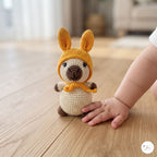 A crocheted kangaroo toy sits on a light wood floor, with a baby's hand and arm resting beside it. The kangaroo is cream-colored with a mustard yellow hood and matching scarf, and dark brown accents on its nose, paws, and ears.