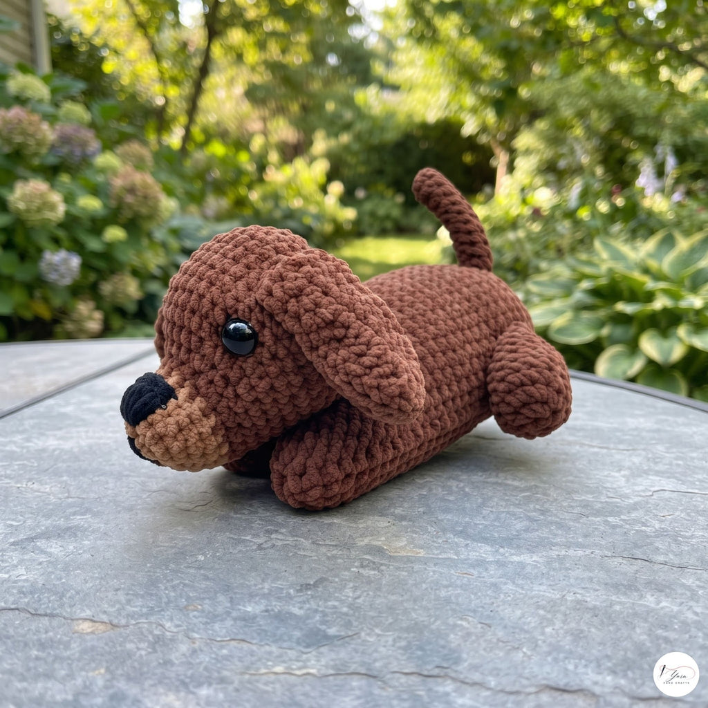 A small crocheted dachshund toy lying on a gray stone table outdoors, with a blurred garden background of green foliage and flowers.